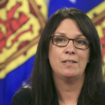 A brunette woman with glasses speaks in front of Nova Scotia flags