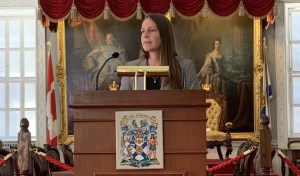 A woman with long brown hair and wearing a grey blazer stands at a podium in a fancy building with a painting of kings and queens in the background.