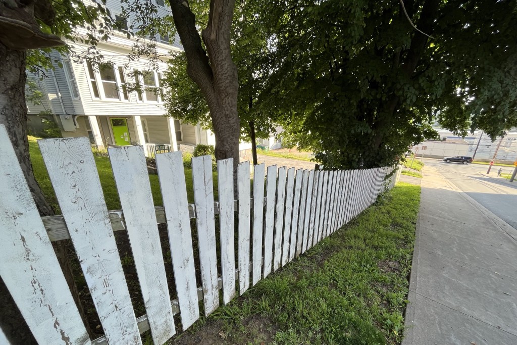 A white picket fence lines a sidewalk. In the background, a vehicle drives on a street perpendicular. It's a sunny day.