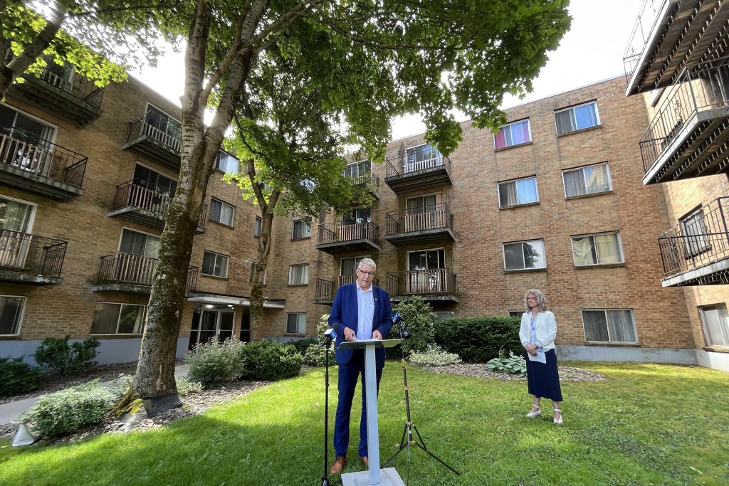 A man in a suit wearing glasses speaks at a podium outside an apartment building on a sunny day. A woman stands behind him.