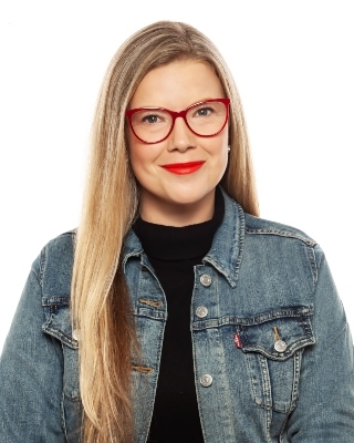 A woman with long blonde hair, red frame glasses, bright red lipstick, and wearing a denim jacket with a black top underneath.