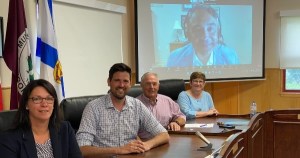Four people sit at a table looking at the camera. In the background is a man on a video screen