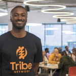 a young Black man wearing a T shirt with the word "Tribe" on it, in a busy conference room