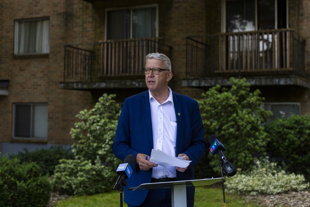 A man in a suit wearing glasses speaks at a podium outside an apartment building on a sunny day.