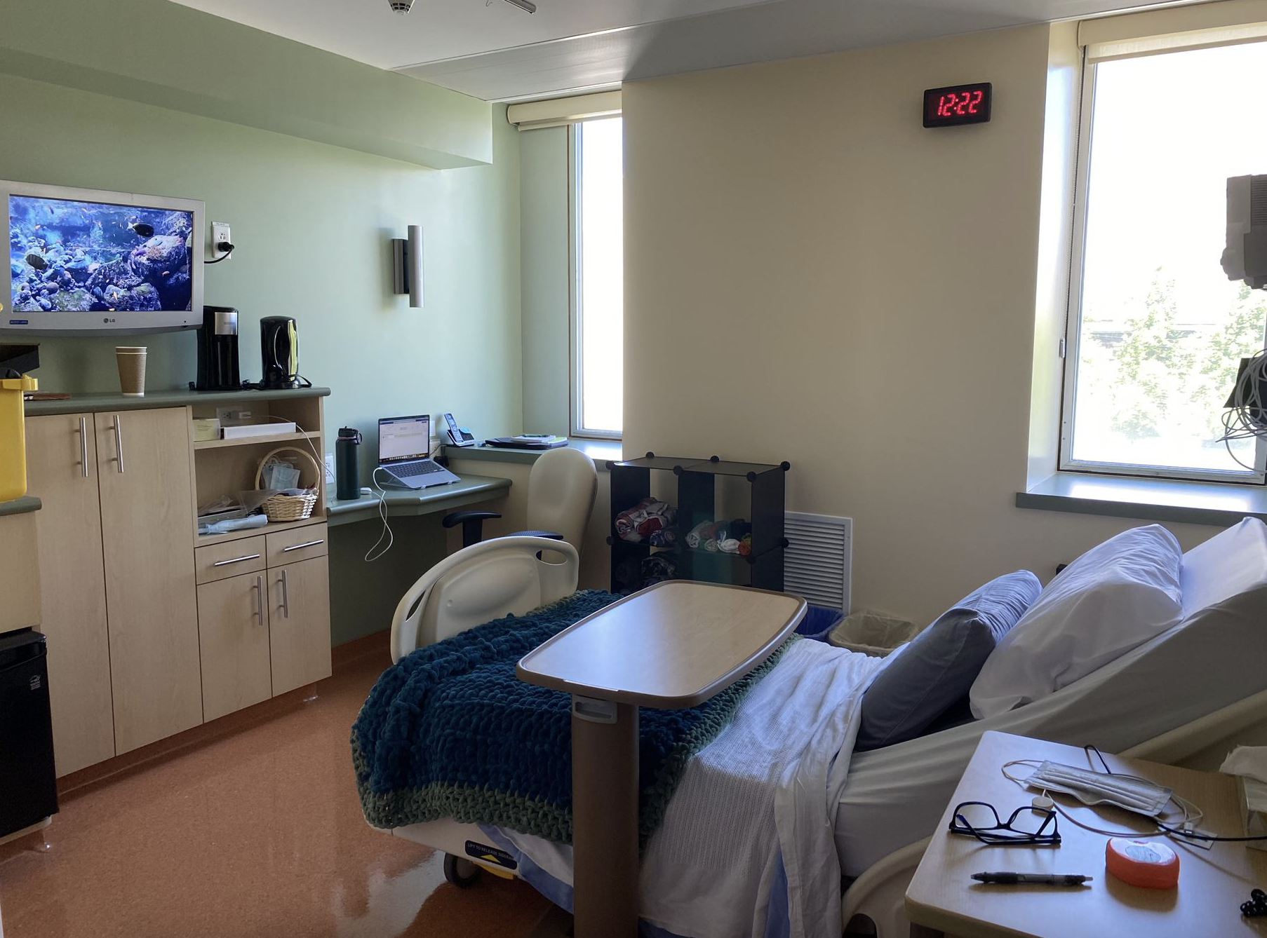 A hospital room with a bed, TV on with screen saver and various items like eyeglasses, cups, and headphones showing someone is inhabiting the room.