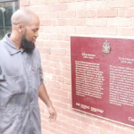 A Black man in grey coveralls admires a red plaque on a brick building