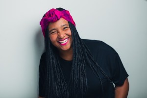 A Black woman with long braids and wearing a bright pink head wrap and black top smile for the camera.