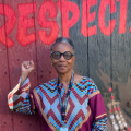 A Black woman with short hair and round black framed glasses, wearing a multi coloured dashiki. She is standing with her right fist raised, in front of a mural of Arethan Franklin with the word 'Respect' in big red letters.